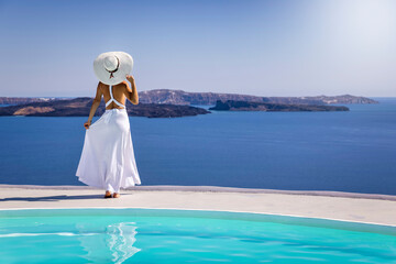 A beautiful woman in a white summer dress stands by the pool and enjoys the breathtaking view over...