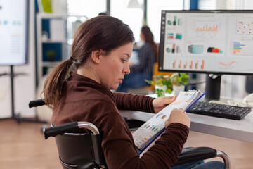 Business woman with disabilities working for deadline project reading documents from clipboard checking graphs sitting immobilized paralized in wheelchair in business office looking on computer.