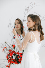 A beautiful young girl with brown hair in a white wedding dress with sleeves. A bride with her hair down in a photo studio looks in the mirror. the decor is made of red flowers. 