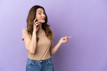 Middle aged caucasian woman using mobile phone isolated on purple background pointing finger to the side