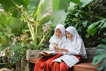 Naklejka na ściany i meble two children wearing veil in school uniforms using a cellphone and a book while studying together sitting on a bench in the park