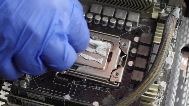 Electronics repair technician in a blue glove thoroughly lubricates the computer processor on the motherboard of a desktop computer with coolant with a cotton swab. Close up