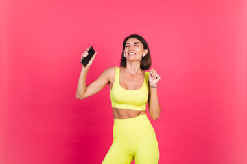 Beautiful fit woman in yellow bright fitting sportswear on pink background happy dancing headphones in ears hold phone listen music excited perfect body