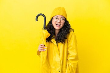 Asian woman with rainproof coat and umbrella isolated on yellow background with surprise facial expression