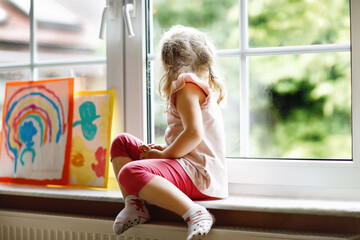 Cute little toddler girl by window with rainbow painted with colorful colors. Preschool on bad...