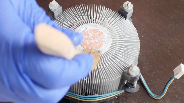 Electronics repair technician in a blue glove sweeps dust off an aluminum heatsink of the computer processor on the motherboard of a desktop computer from old coolant with a special brush. Close up