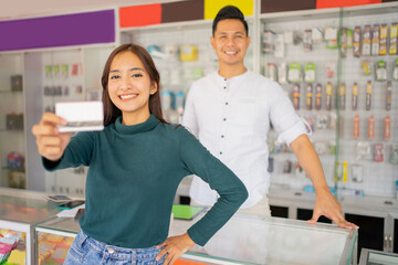 Obraz premium a female customer smiles while showing a credit card for cashless payment to the camera with a man in the background in a smartphone shop