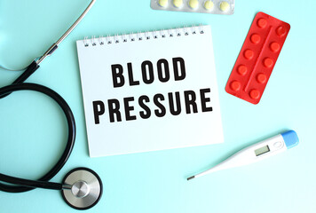The text BLOOD PRESSURE is written on a white notepad that lies next to the stethoscope and pills on a blue background.