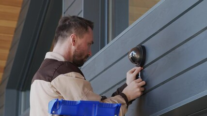 Slowmo tracking shot of handyman in uniform standing on ladder and using screwdriver to install home security camera on exterior of house