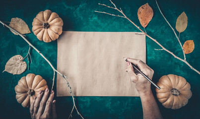 Halloween grunge dark background, pumpkins, dry branches, orange leaves decorations. Autumn composition frame, party invitation, flat lay, vintage paper, woman hand with pencil, copy space.