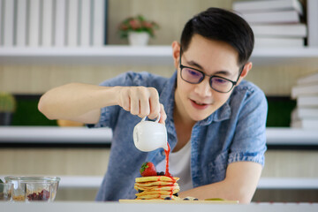 in modern home, young man with glasses enjoy to practice his chef skill by decorating great healthy breakfast by slow pouring sweet strawberry jam onto tower of tasty pancakes