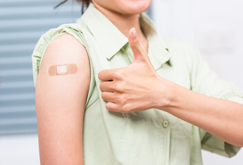 Asian young woman showing gesturing thumb up or like and her arm with plaster band aid after coronavirus Covid-19 vaccine injection on shoulder, coronavirus COVID-19 vaccination concept