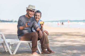 Happy senior couple smile sitting chair beach relaxing outdoors hold and showing credit card...