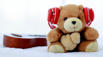 in home bedroom, brown lovely fluffy teddy bear toy creatively wearing red earphone and sit on bed near guitar as lovely doll for enjoy music playing on delight celebration holiday