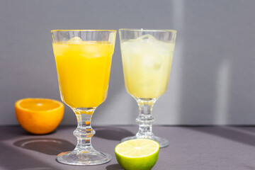 a pair of glasses with a cold citrus drink or lemonade or alcoholic cocktail on gray background, two vintage goblets among sliced orange and lime, copy space, minimalism