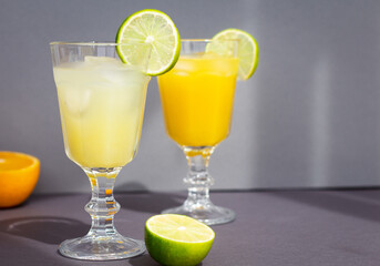 two glasses with a cold citrus drink or lemonade or alcoholic cocktail on gray background, a pair of vintage goblets among sliced orange and lime, copy space