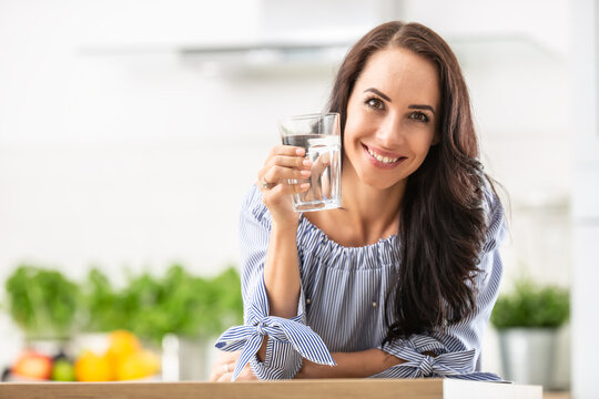 Smiling pretty woman holds a glass of water leaning on kitchen desk