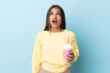 Young Uruguayan woman with strawberry milkshake isolated on blue background looking up and with surprised expression