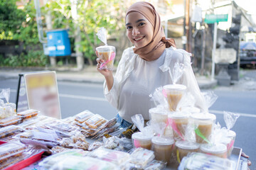 A girl in a veil holds takjil food filled with kolak in a plastic cup at a roadside stall