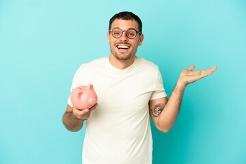 Brazilian man holding a piggybank over isolated blue background with shocked facial expression