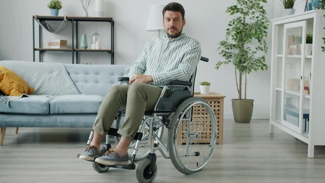 Portrait of man with physical disability sitting in wheelchair in apartment looking at camera indoors in comfortable house. People and health concept.