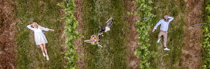 aerial drone shot, pic of blonde woman with white dress and her boyfriend or husband posing with bicycle with beautifully decorated flower basket in nature, nature concept, flowers, delivery service