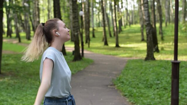 Young woman wearing earphones is dancing and listening to music in a city park in summer. Positive energy