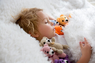 Little toddler child, cute blond boy, sleeping with many teddy bears, handmade amigurumi toys at home