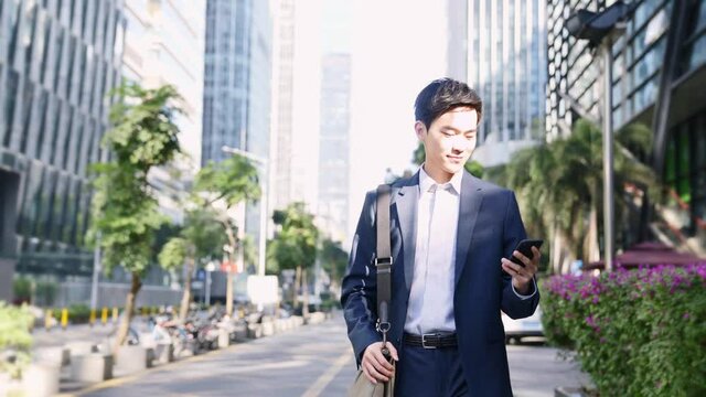 young asian business man commuter looking at cellphone while walking on street in downtown financial district of modern city