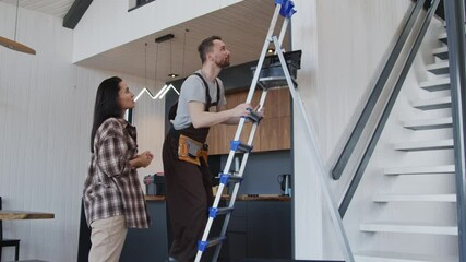 Slowmo handheld tracking shot of young female homeowner smiling and talking to handyman He is getting up on ladder and fixing smart home device or sensor - Powered by Adobe