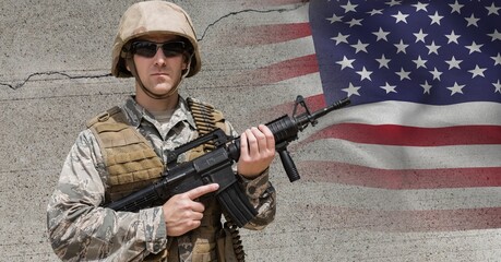 Composition of male soldier wearing sunglasses holding gun, against concrete wall and american flag