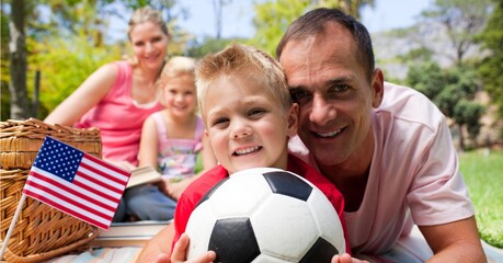 Composition of waving american flag, over happy family with football having picnic