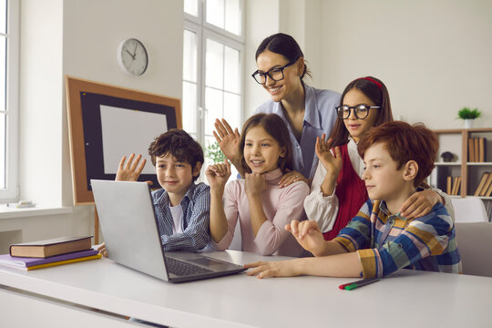 Teacher and elementary student children using laptop for online video call sitting at classroom desk. happy smiling young adult people waving hands with greeting gesture looking at screen