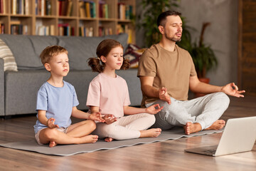 Dad with child little son and daughter doing yoga online, sitting in lotus pose on floor and meditating with closed eyes