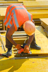 A worker in a protective helmet saws off a wooden plank at a construction site - obrazy, fototapety, plakaty