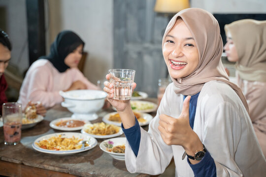 a veiled woman smiles at the camera with thumbs up while holding a glass for breaking fast together in the dining room