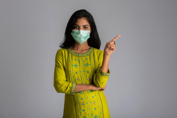 Young girl wearing a medical face mask posing, pointing and showing something on grey background.