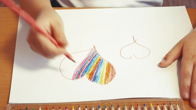 A rainbow heart is drawn by children's hands in close-up with a colored pencil. The concept of children's creativity, online education. Leisure of a happy child, quarantine and isolation. View from