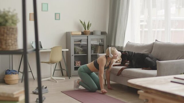 Wide shot of blonde Caucasian woman wearing headphones, leggings and sports top, standing on knees on floor at home, kissing and petting her big Doberman dog lying on couch