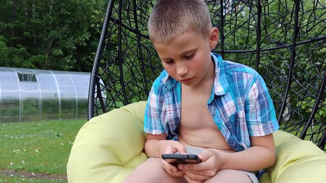 Boy in plaid shirt sitting in cocoon chair and playing with telephone. Smartphone games. Summer time