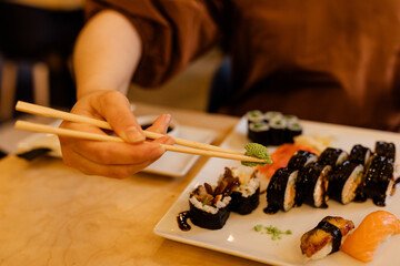 Obraz premium Women's hands skillfully use Chopsticks at table in a restaurant. An unrecognizable person uses wasabi for sushi