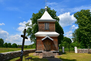 A close up on an old wooden Orthodox shrine located next to a cemetery with a tall stone wall covering all sides of it seen next to a dense forest or moor and a field in the middle of a Polish village