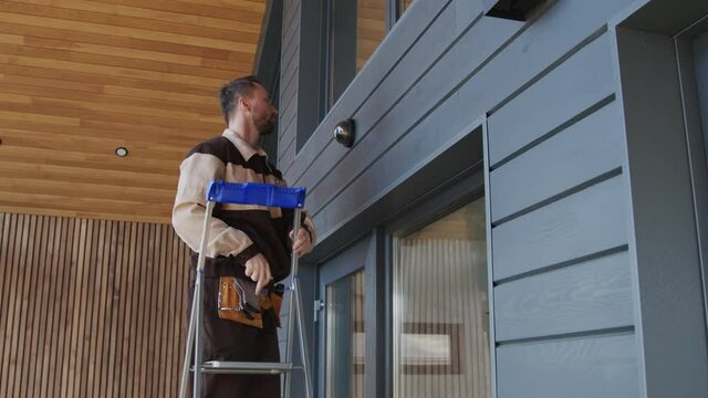 Handheld slowmo tracking shot of male contractor in uniform getting up on ladder and installing home security camera on exterior of clients house