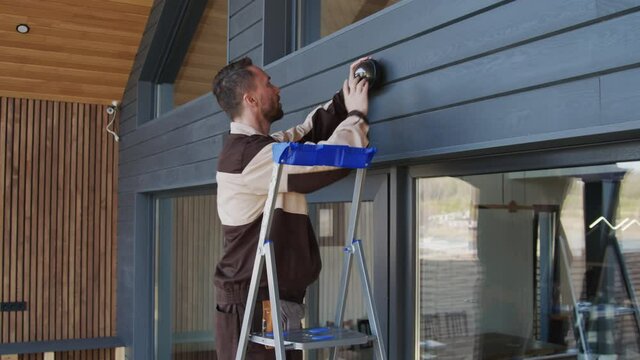 Slowmo tracking shot of handyman standing on ladder and installing home security camera on exterior of house