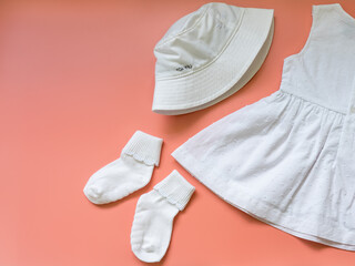 A set of summer clothes for kids on a pink background. White baby dress, Panama, and socks