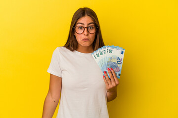 Young caucasian woman holding bills isolated on yellow background shrugs shoulders and open eyes confused.