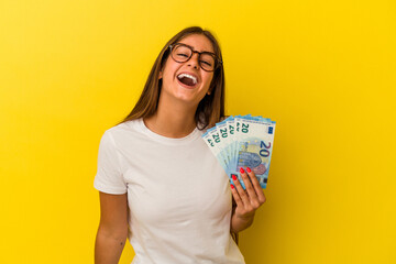 Young caucasian woman holding bills isolated on yellow background laughing and having fun.