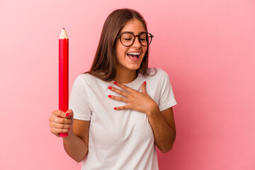 Young caucasian woman holding a big pencil isolated on pink background laughs out loudly keeping hand on chest.