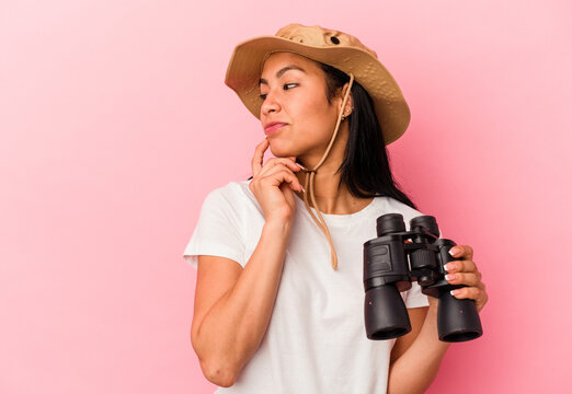 Young mixed race explorer woman holding binoculars isolated on pink background relaxed thinking about something looking at a copy space.