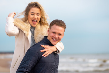 Happy man giving piggyback ride to his woman and laughing at beach. Smiling guy in love carrying on...